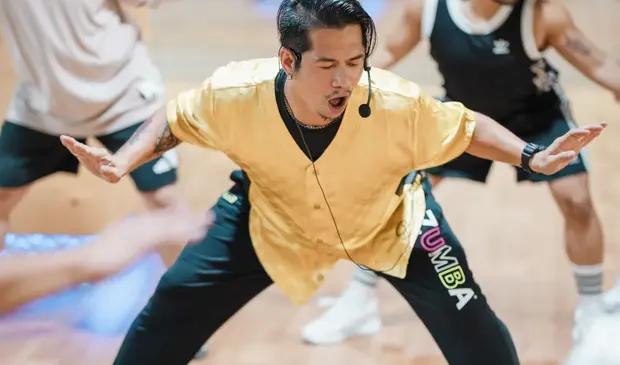 Energetic BODYJAM instructor in a yellow shirt leading a lively dance routine at GymNation, surrounded by motivated participants following his moves in a vibrant fitness studio.
