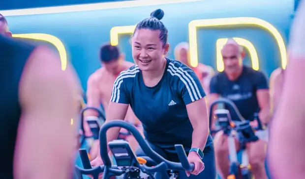 Smiling woman in a LES MILLS SPRINT class at GymNation, riding an indoor bike with neon lights in the background.