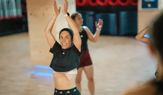 A smiling woman raising her arms while dancing during a belly dancing fitness class at GymNation, surrounded by other participants