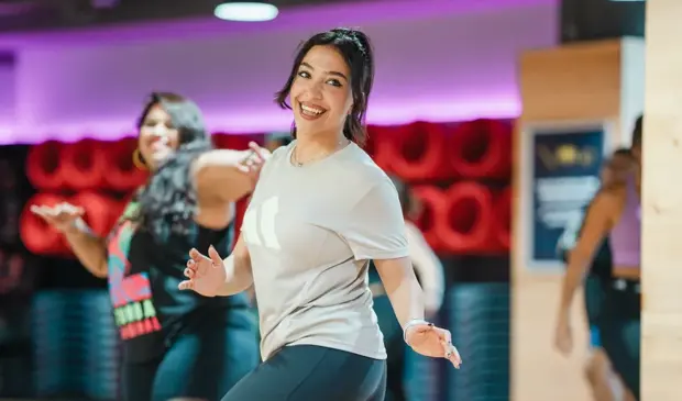 A cheerful woman mid-step during a belly dancing fitness class at GymNation, surrounded by smiling participants