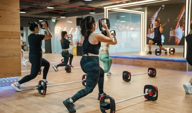 Members using Les Mills SMARTBAR™ equipment during a BODYPUMP class at GymNation, following the instructor on stage inside a fitness studio.