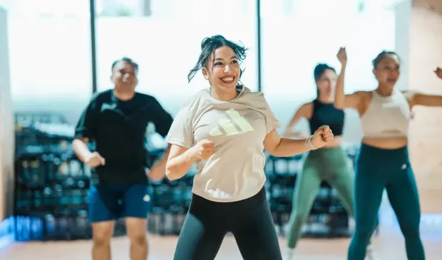 A group of smiling participants dancing during a Zumba class at GymNation, with a female participant leading front and center in a bright, energetic studio.