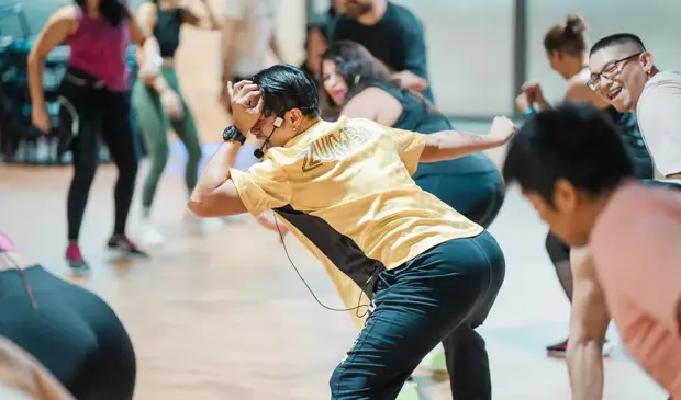 Zumba instructor in a yellow shirt leading a lively mixed-group class at GymNation, with participants dancing, smiling, and enjoying the atmosphere.