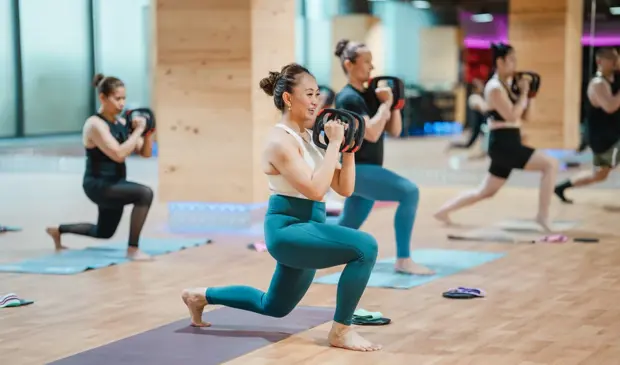 Participants holding weighted plates while performing lunges during a Mat Pilates class at GymNation, focusing on full-body strength, stability, and controlled breathing.