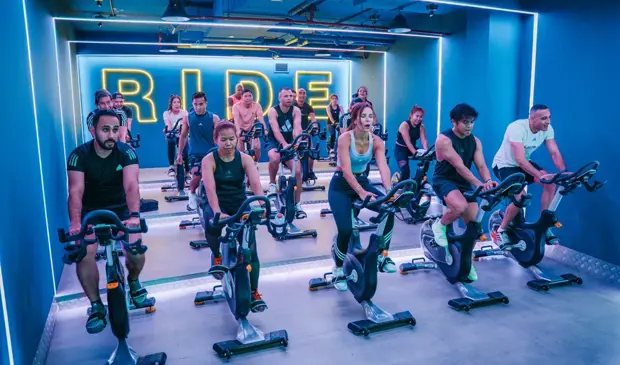 People cycling in a high-energy LES MILLS SPRINT class at GymNation, with neon lights and “RIDE” sign in the background.
