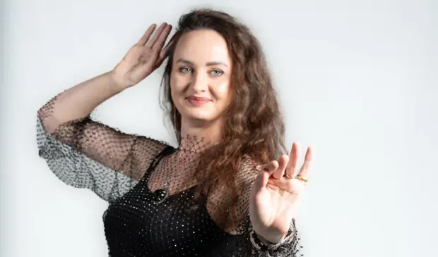 A professional belly dance instructor posing gracefully in a sparkling black outfit with sequined hip scarf, smiling confidently against a white background.